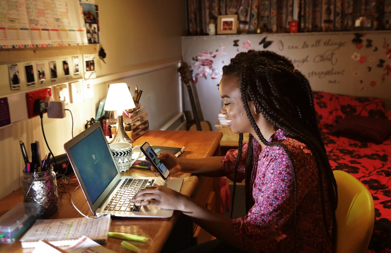 College-age female sits at her desk in her dorm room. Her laptop is open in front of her, and she is looking at her phone, smiling. 
