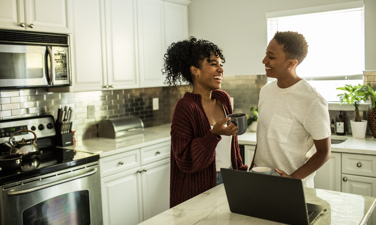 Two women smile at each other while in their kitchen. One is holding a cup of coffee, while the other has her laptop open in front of her. 