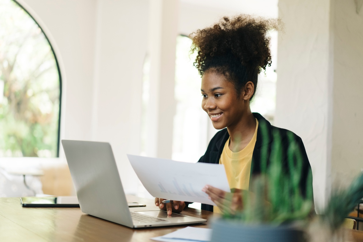 A woman smiles while looking at her laptop at home while holding a piece of paper