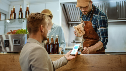 A man working behind the counter at a coffee shop holds up a payment processing device a customer uses to pay using a digital wallet on his phone.