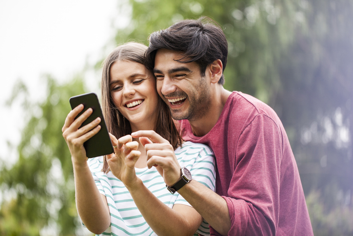 Happy young couple look at their savings account on their smartphone after comparing credit union savings vs. bank savings accounts.