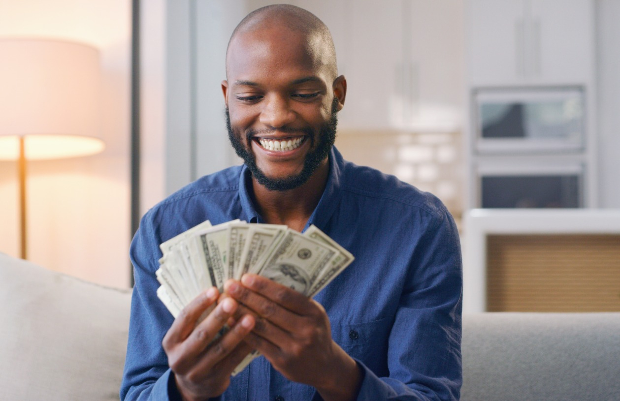 A man sit on a couch holding and looking at many dollar bills in his hands.