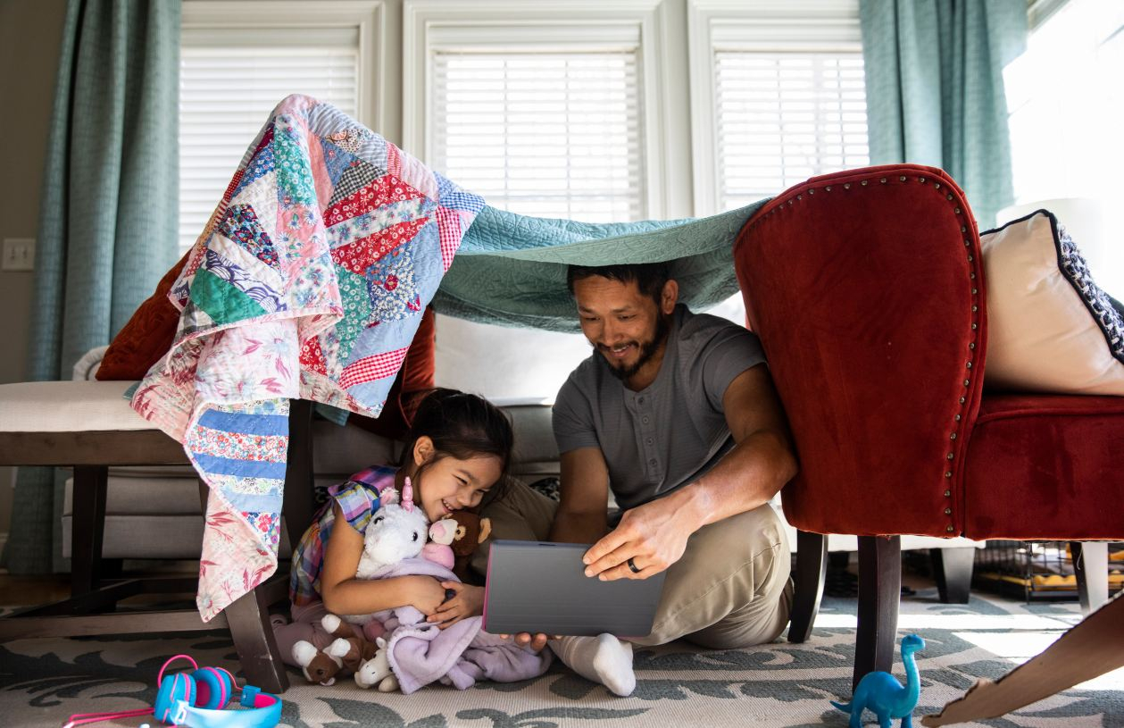 A father and his daughter use a tablet under a blanket fort