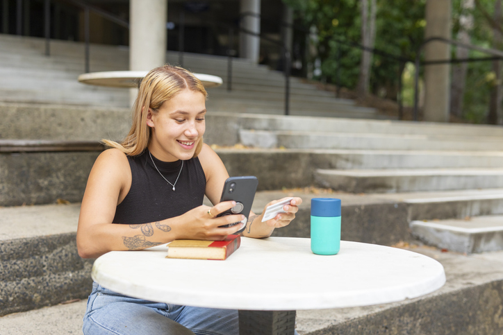 College-age young woman happily makes a purchase on her first credit card while sitting outside on campus.