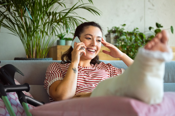 Young woman with broken leg happily tells friend on phone that her expenses were covered by her emergency fund in her high-yield savings account.