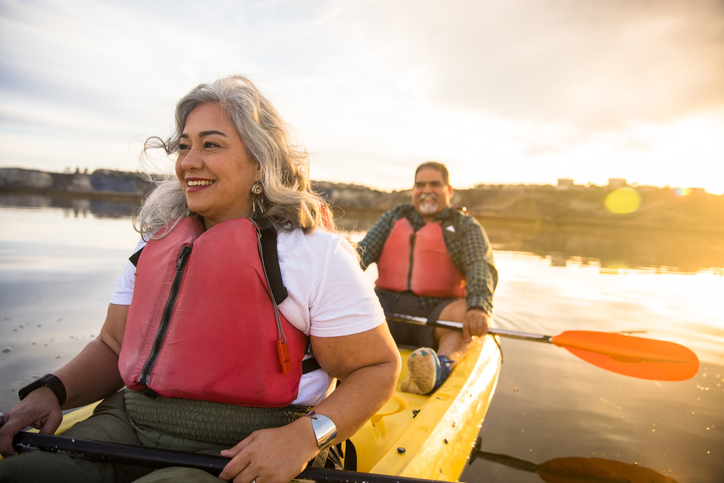 Senior Latinx couple happily kayaks at sunset knowing they navigated retirement planning with help of certificates.