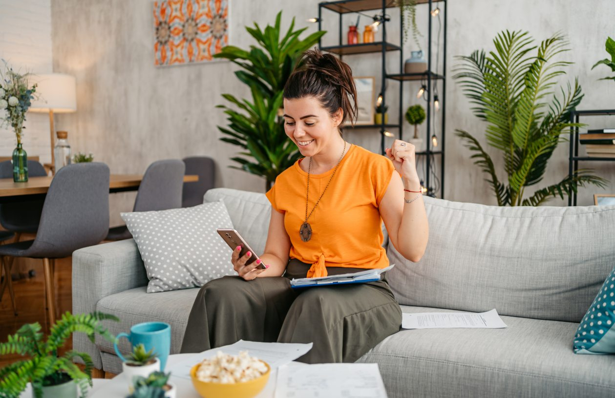 A woman sits on a couch on her phone with an expression of happiness and excitement as she pumps her fist in the air
