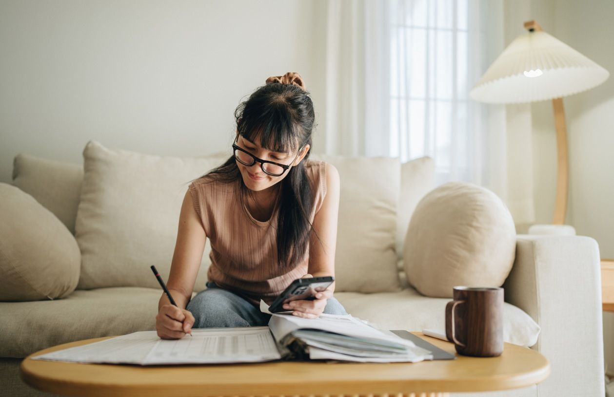 A woman sits on a couch in her home and writes in a notebook while holding her phone