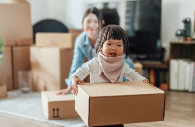 Toddler girl playfully moves boxes into new home as mother smiles in distance. 