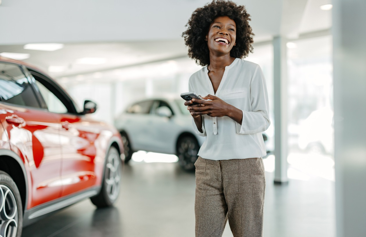 A woman holds her phone at a car dealership while looking off to the side and smiling
