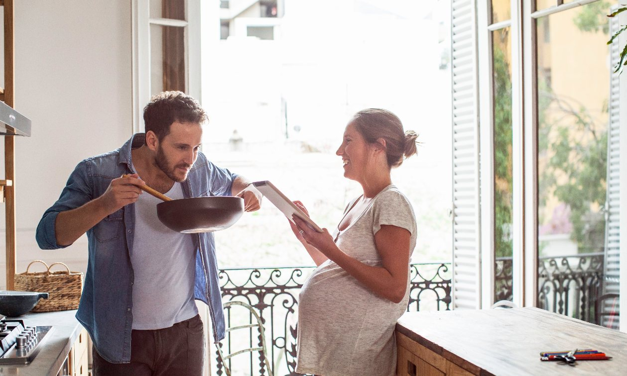 Husband and pregnant wife cook in the kitchen. The soon-to-be mom reads a book and smiles, as the husband smells the dinner he is making. 