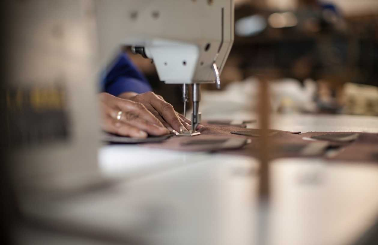 A woman's hands run a piece of fabric through a sewing machine that sits on a clothing factory table.