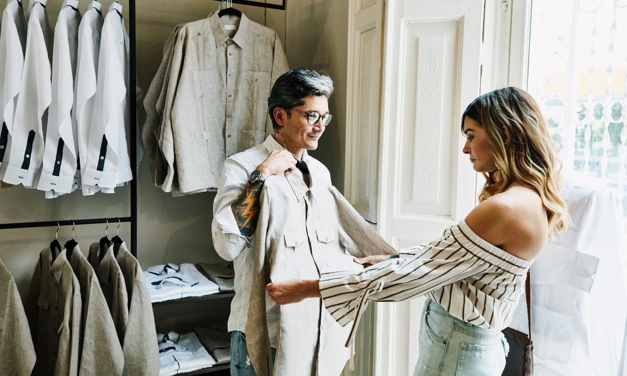 In a clothing store, a mature man holds up a crème blazer, laying it over his chest. A woman he is shopping with tugs at the size of the blazer to measure the fit. 