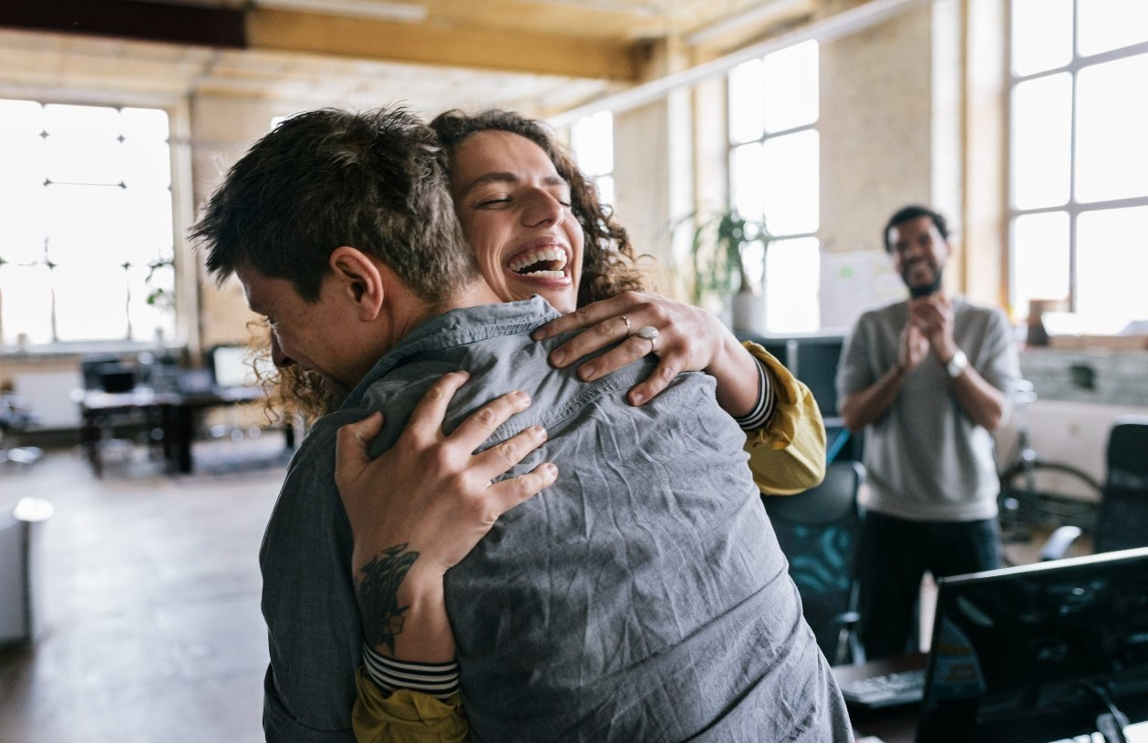 Female employee hugs male employee with a big smile on her face, as other coworkers surround them, clapping in celebration. 