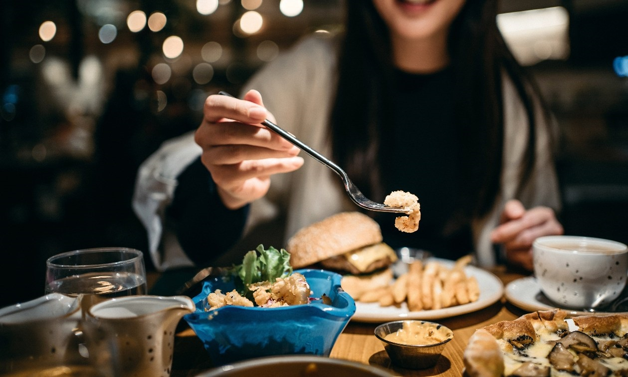 Woman sits at a full table of food inside a restaurant. She brings a fork to her mouth. 
