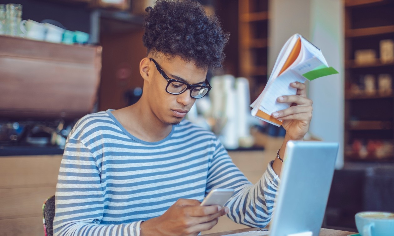 A young man sits at a coffee shop table with his open laptop before him. He looks down at his ringing cell phone in one hand as he holds an open pad of paper in the other. 