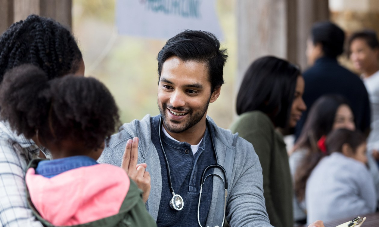 A doctor in street clothes with a stethoscope around his neck holds up two fingers to test the eyesight of a little girl being held by her mother. 