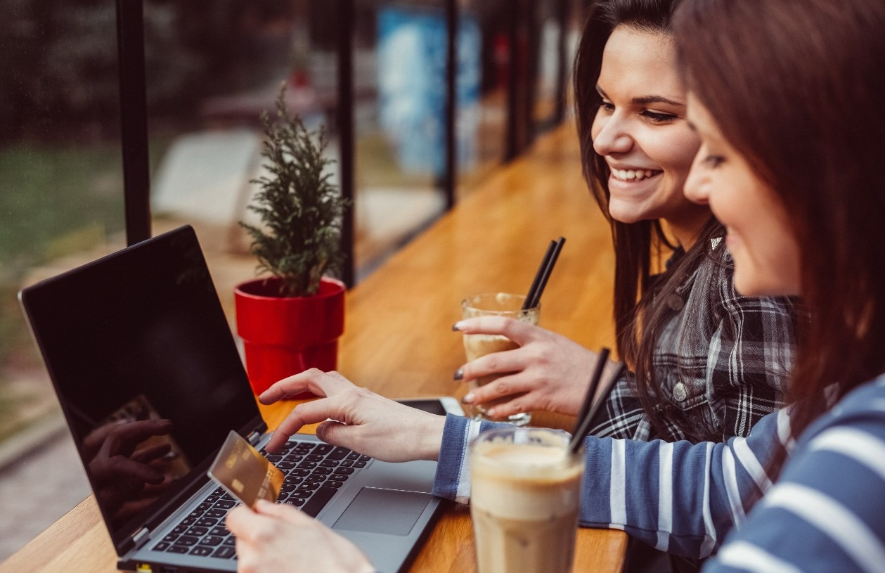 Mother and teenage daughter sit at a countertop at a coffee shop. The teen is holding her credit card while typing on her laptop. 