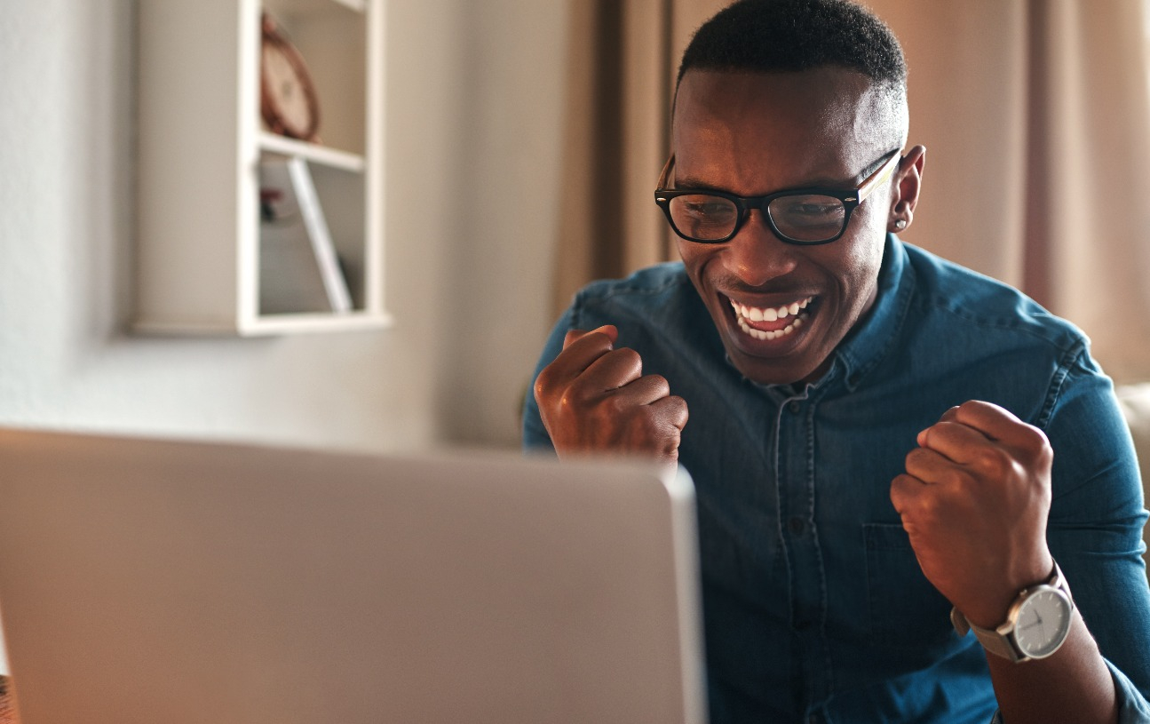 A man is sitting at his desk, cheering as he looks at his laptop screen.